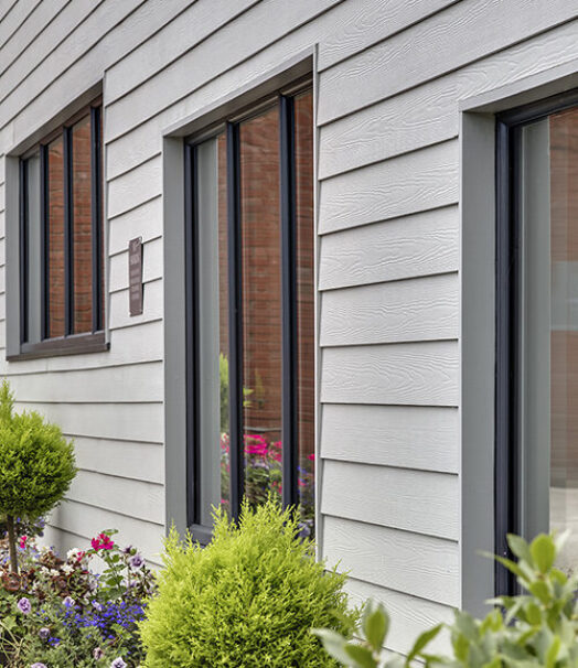 A close-up image of a building facade clad in grey planks with small bushes in front.