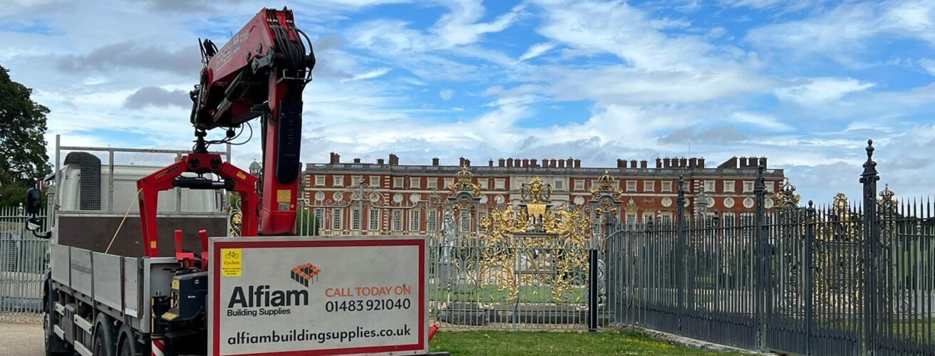 An Alfiam delivery truck parked outside of the golden gates of Hampton Court Palace, under a bright blue sky with a few clouds.