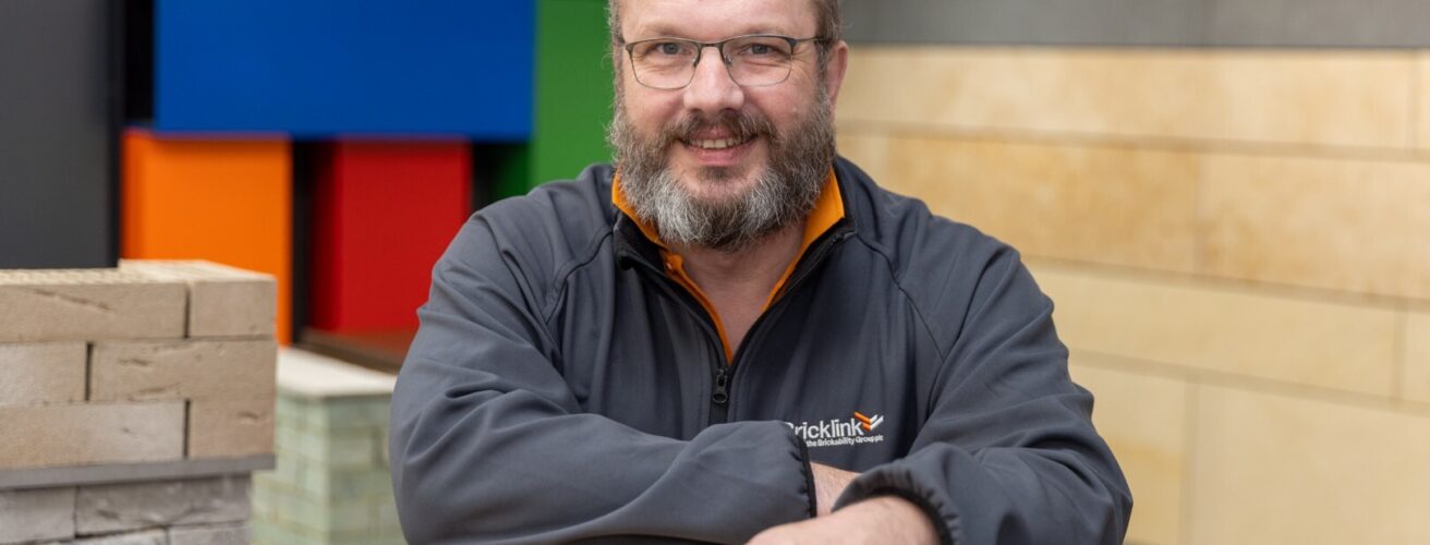 A man stands smiling leaning on a pile of bricks in a construction showroom.