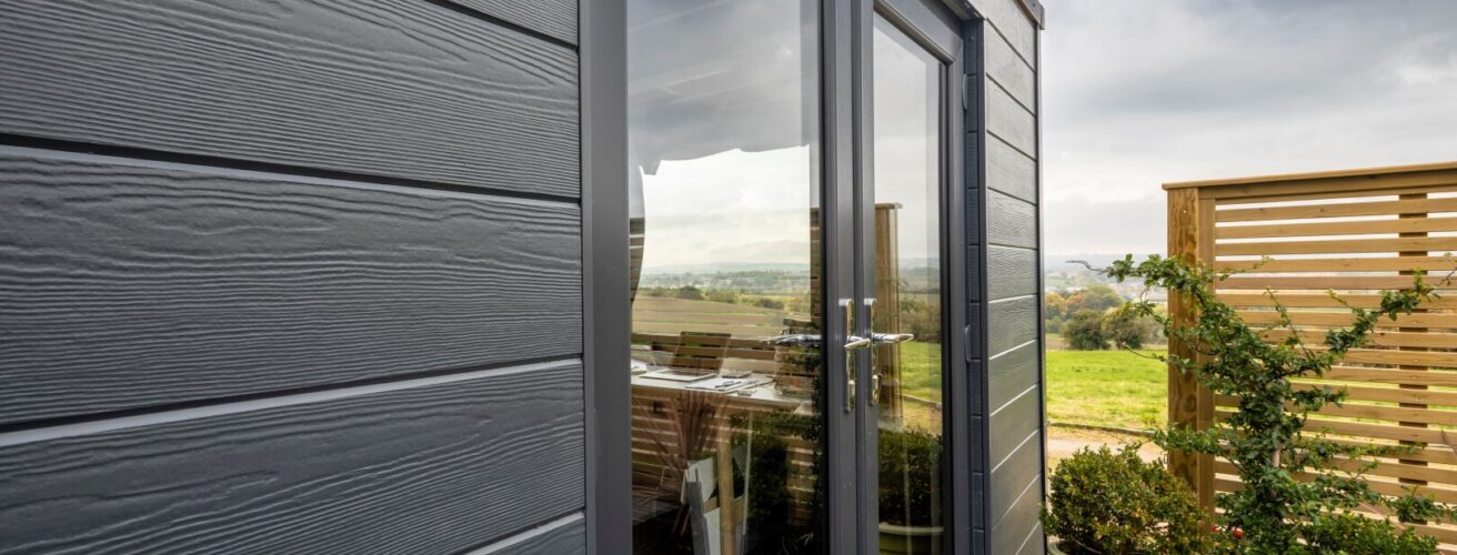 An image depicting an outdoor shed featuring dark plank cladding with a countryside view in the background.