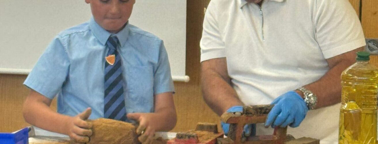 A man and boy stand at the front of a class behind a table, moulding clay into bricks.