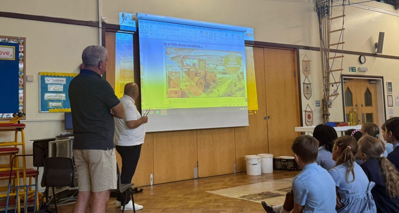 Two adults stand in front of a class of children in a school hall giving a presentation on brick manufacturing.