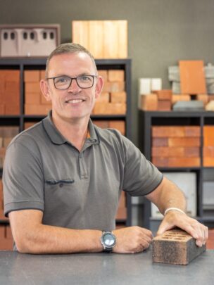 A man stood at a counter holding a brick with a range of bricks displayed on shelves in the background.