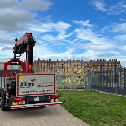 An Alfiam delivery truck parked outside of the golden gates of Hampton Court Palace, under a bright blue sky with a few clouds.