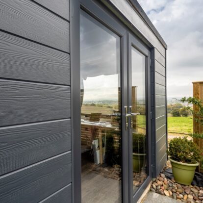 An image depicting an outdoor shed featuring dark plank cladding with a countryside view in the background.