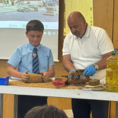 A man and boy stand at the front of a class behind a table, moulding clay into bricks.
