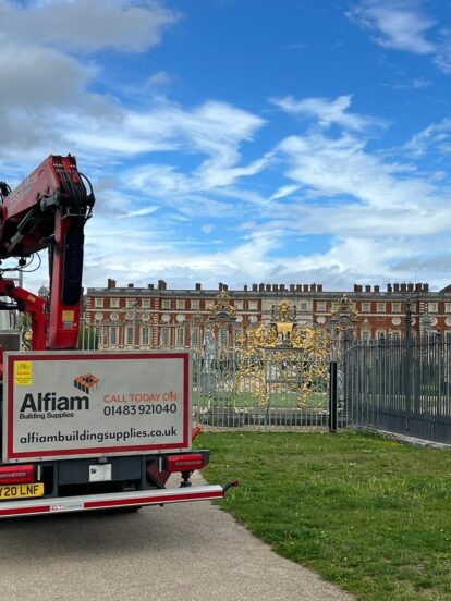 An Alfiam delivery truck parked outside of the golden gates of Hampton Court Palace, under a bright blue sky with a few clouds.