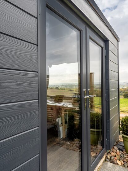 An image depicting an outdoor shed featuring dark plank cladding with a countryside view in the background.