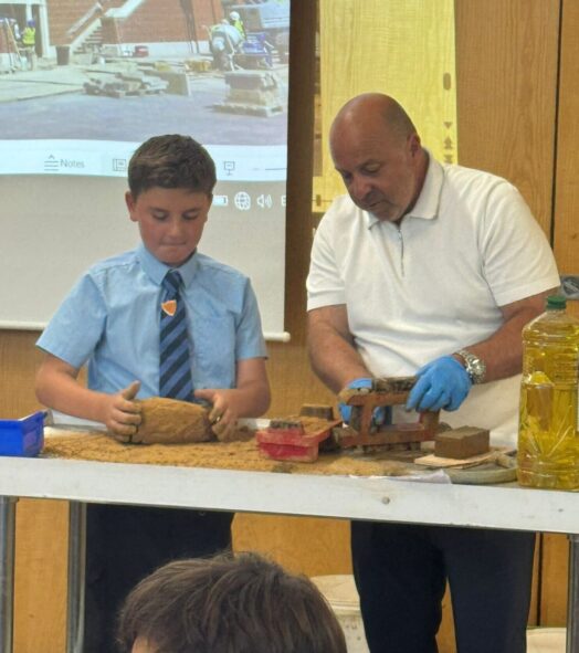 A man and boy stand at the front of a class behind a table, moulding clay into bricks.