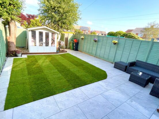 A very neat garden with pale grey porcelain slabs encircling a freshly mowed lawn. there is a shed at the far end of the garden and garden furniture in the front right hand corner.