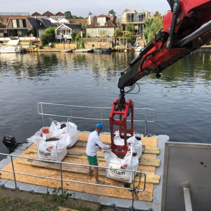 A person helps to guide Alfiam aggregate sacks onto a river barge as they're lowered by a red crane.