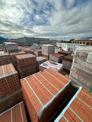 A photograph taken across a large stack of red bricks. A small portacabin is visible in the background.
