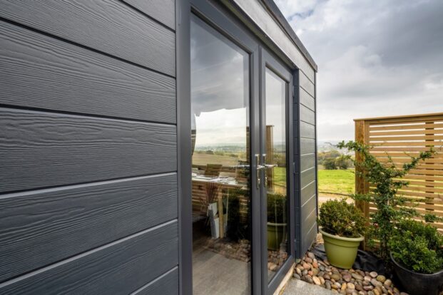 A garden room with dark grey, wood-effect walls, situated in a garden with a view of a slightly cloudy sky.