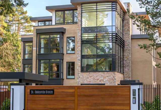 A grand, modern house featuring a stone and brick facade behind a closed wooden gate.