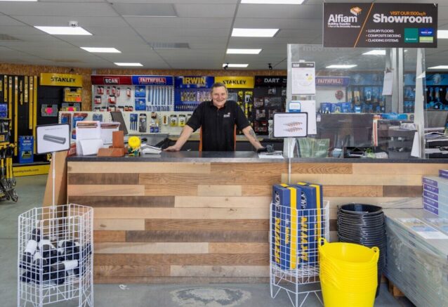 An Alfiam employee standing behind the customer desk and checkout with building materials and tools int he background.