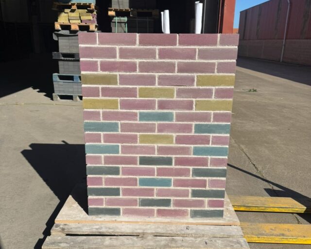 A sample brick wall, sitting on a palette, made from pink, yellow and blue bricks.