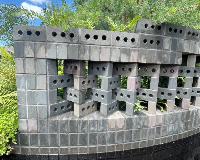 A decorative brick wall in dark grey, with lush greenery in the background and water around the base.