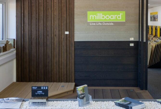 A photo of composite cladding and decking fitted on a showroom wall and floor, with a green sign reading 'Millboard, Live.Life.Outdoors' mounted on the wall.