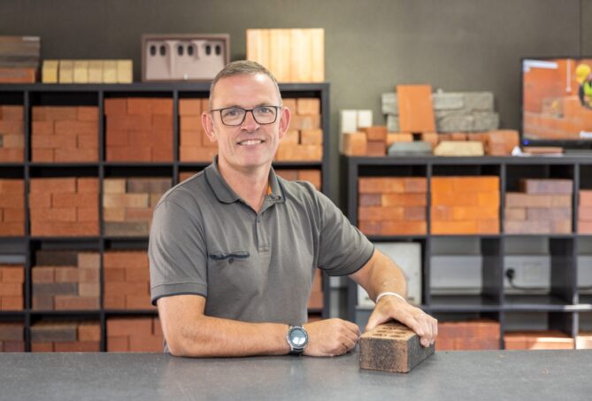 A man stood at a counter holding a brick with a range of bricks displayed on shelves in the background.