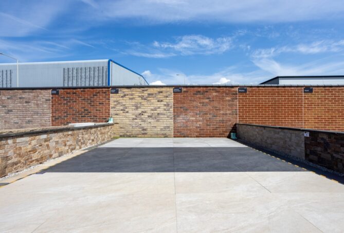 An image of neatly laid porcelain paving in various shades of light and dark grey, with a brick wall framing the space, on a sunny day.