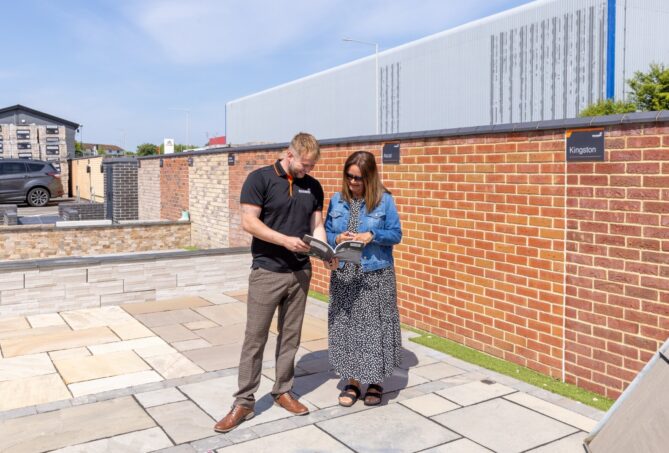 Two people stood among a paving display, surrounded by brick walls, looking at a brochure under a clear sky.