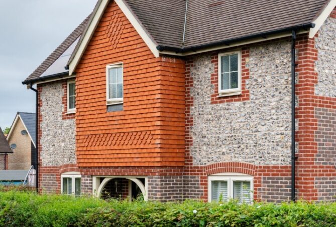 The front of a house with a flint block facade, partially obstructed by a hedge.