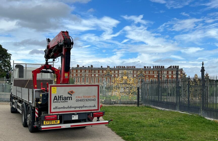 An Alfiam delivery truck parked outside of the golden gates of Hampton Court Palace, under a bright blue sky with a few clouds.