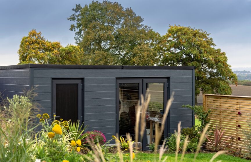 An image of a small cabin in a back garden, clad in a dark grey Hardie Plank boards, surrounded by greenery.